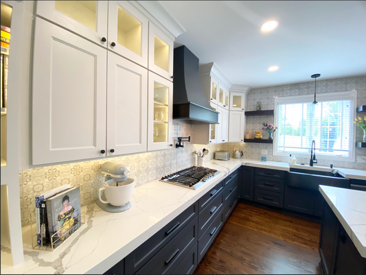 white top cabinets with blue bottom cabinets, white uppers and navy base, black hardware, pattern backsplash, wood hood, recessed lighting, window, wood floor, glass cabintry.